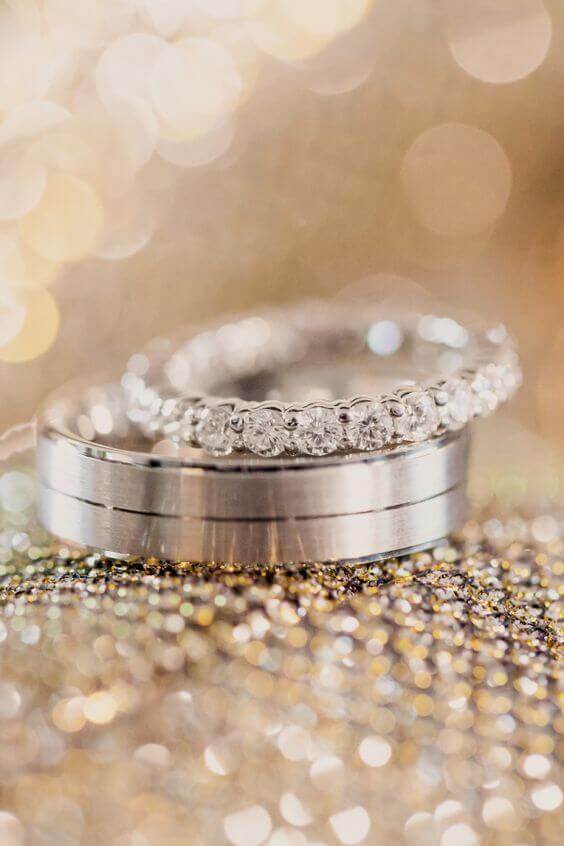 Diamond eternity ring stacked on a brushed platinum wedding band, glimmering on a sequined champagne backdrop with warm bokeh.