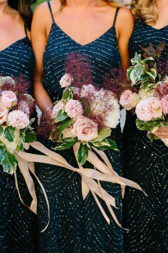 Three bridesmaids in navy beaded slip dresses hold blush-and-mauve bouquets with cascading champagne ribbons, embodying a romantic evening wedding.