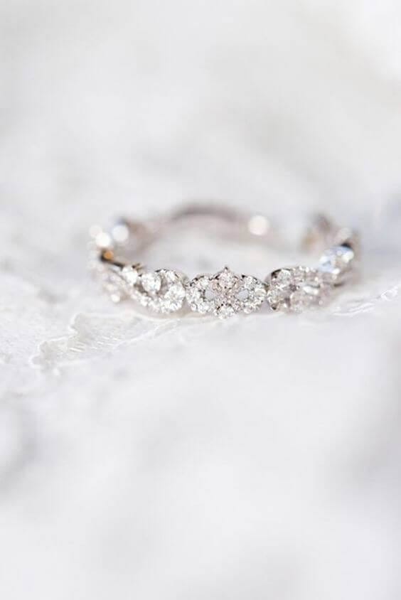 Close-up of a diamond eternity wedding band featuring marquise and round clusters, resting on delicate white lace with soft bokeh.