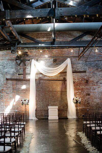 34 budget wedding ideas for a creative affordable wedding 8 Minimalist wedding ceremony in an industrial loft, featuring a white chiffon draped arch, floral arrangements, and wooden chairs with scattered petals.