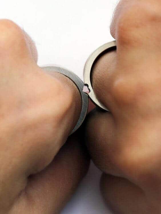 Two silver-toned wedding bands on adjacent fingers press together to form a heart shape against a clean white background.