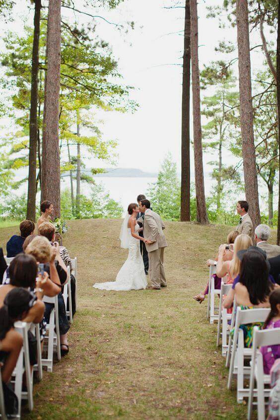 34 budget wedding ideas for a creative affordable wedding 9 Bride and groom kiss in a serene lakeside pine grove, surrounded by guests in white chairs, creating a romantic budget wedding moment.