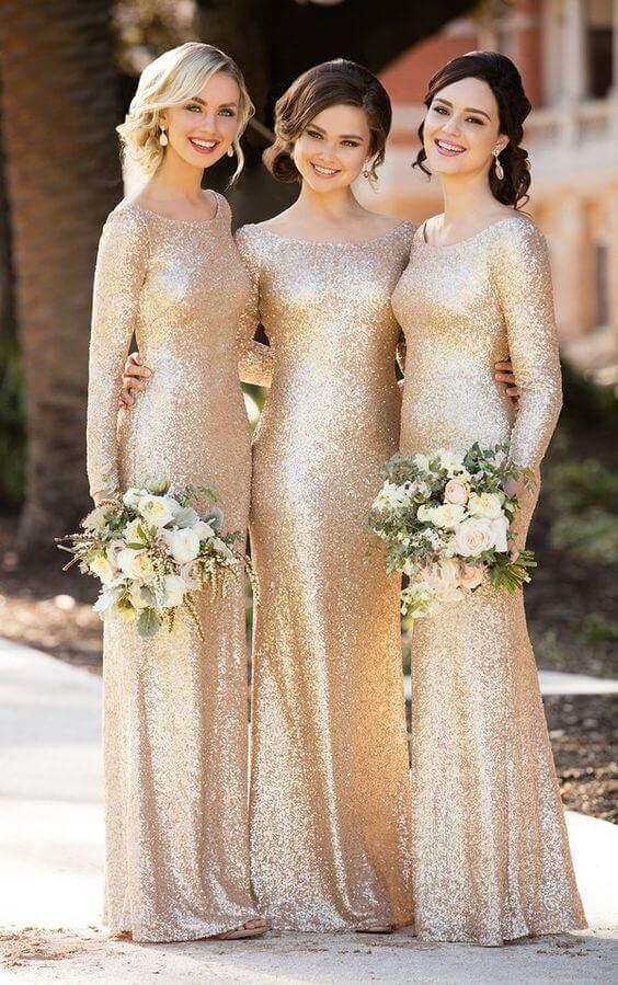 Three bridesmaids in champagne-gold sequin gowns with blush-ivory bouquets stand arm-in-arm on a sunlit garden path, exuding modern glam.