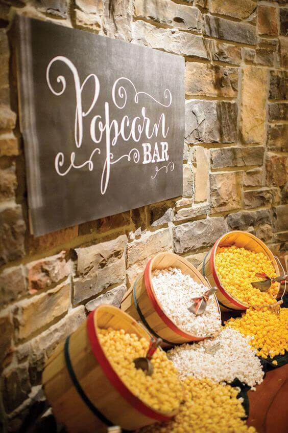 34 budget wedding ideas for a creative affordable wedding 10 Rustic popcorn bar with a chalkboard sign, wooden baskets of buttery yellow and white popcorn, set against a stone wall for a cozy wedding snack.