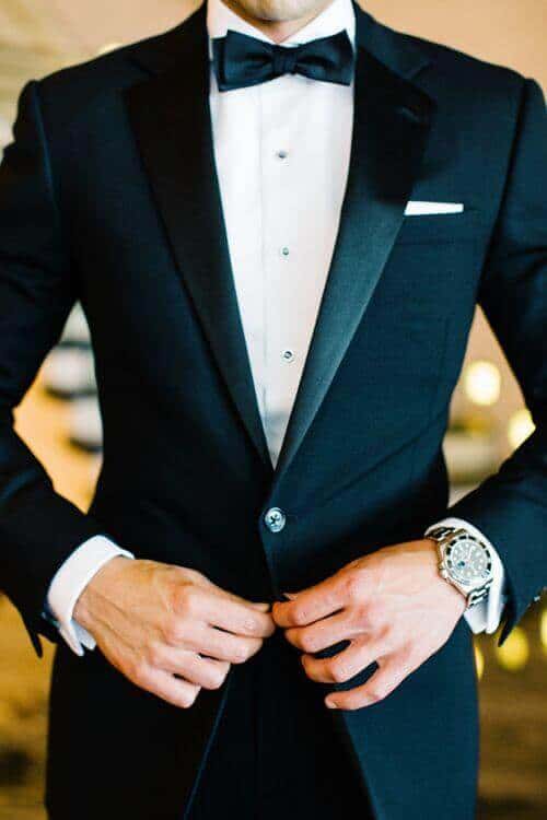 38 wedding accessories list - ideas to transform your big day into magic 10 Close-up of a groom buttoning his tailored black tuxedo, showcasing a crisp white shirt, black bow tie, and elegant details under warm lighting.
