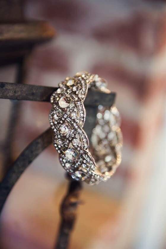 {{post_main_keyword}} crystal bridal bracelet with a braided design, resting on a dark iron stand against a softly blurred brick background.