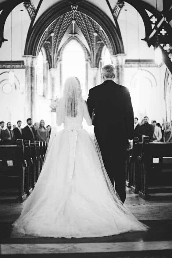 31 steps wedding planning checklist 10 A bride in a full ballgown with a cathedral-length train walks down the aisle of a grand gothic church, escorted by a suited companion.