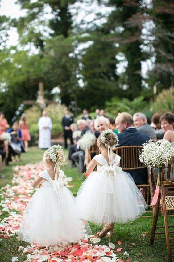 43 cute and perfect flower girl dresses that make every little princess shine 11 Two flower girls in ivory tulle dresses with oversized satin bows walk down a petal-strewn aisle, surrounded by guests and lush greenery.