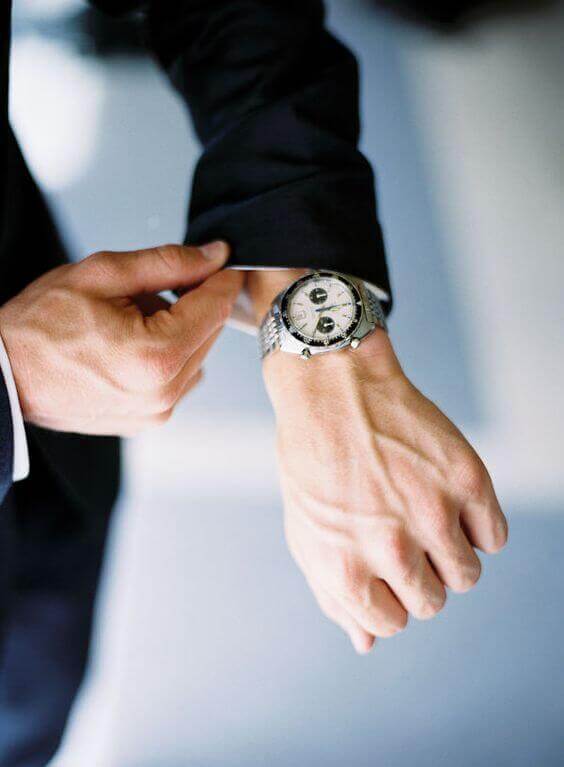 38 wedding accessories list - ideas to transform your big day into magic 11 Refined close-up of a groom's hands adjusting a black suit cuff, showcasing a polished stainless-steel chronograph watch, symbolizing elegance in wedding accessories.