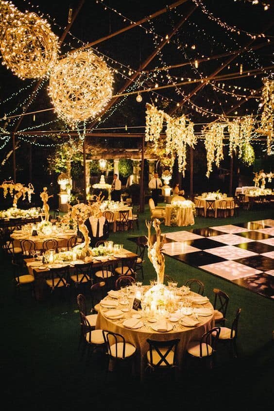 Warmly lit outdoor night wedding reception under twinkling string lights and floral chandeliers, featuring round tables and a black-and-white dance floor.
