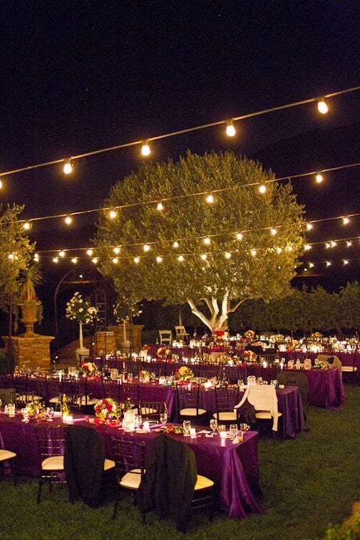 Romantic outdoor night wedding setup with illuminated olive tree, deep purple linens, candlelit tables, and lush floral centerpieces.