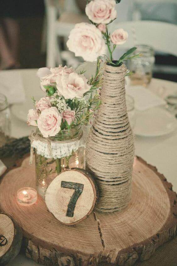 Rustic burlap wedding centerpiece featuring a twine-wrapped bottle with blush roses, a mason jar of florals, and a wood-slice table number.