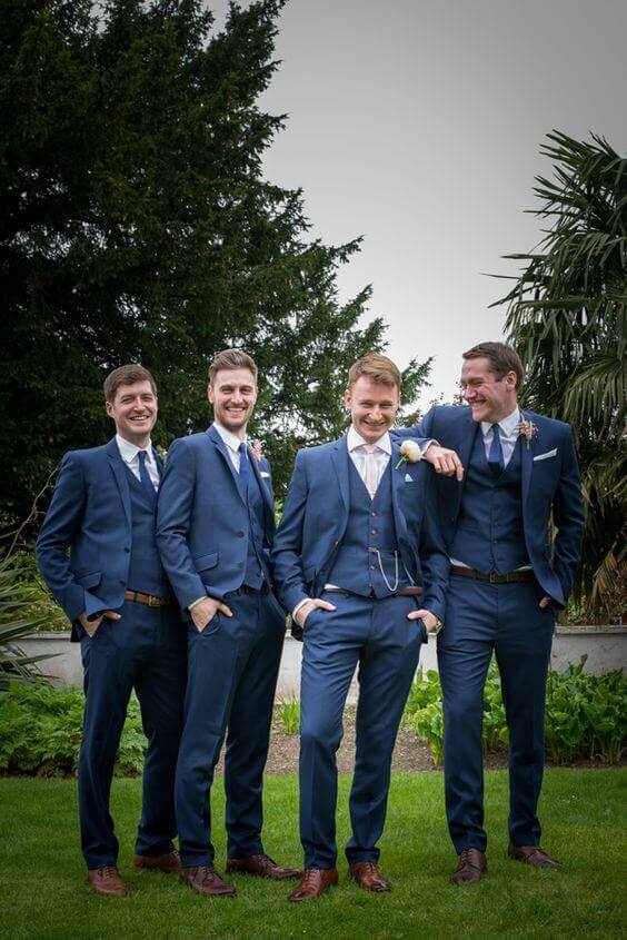 31 amazing wedding groomsmen suits suggestions 13 Four men in coordinated navy three-piece suits with light blue ties stand casually on a manicured lawn, smiling against lush greenery.