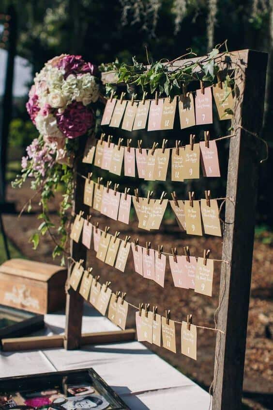 Rustic outdoor wedding seating chart with kraft and blush escort cards clipped to twine on a wooden frame, adorned with hydrangeas and ivy.