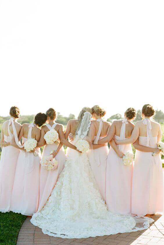 37 Morning Wedding Ideas filled with Sunshine color pallets 14 Bride in an ivory lace gown with a cathedral veil stands with seven bridesmaids in blush dresses, facing a sunlit garden horizon.