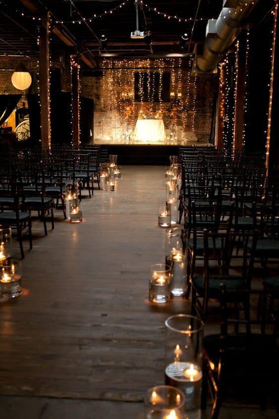 Candlelit aisle in an industrial loft, leading to a softly lit altar against exposed brick, creating a romantic night wedding ambiance.
