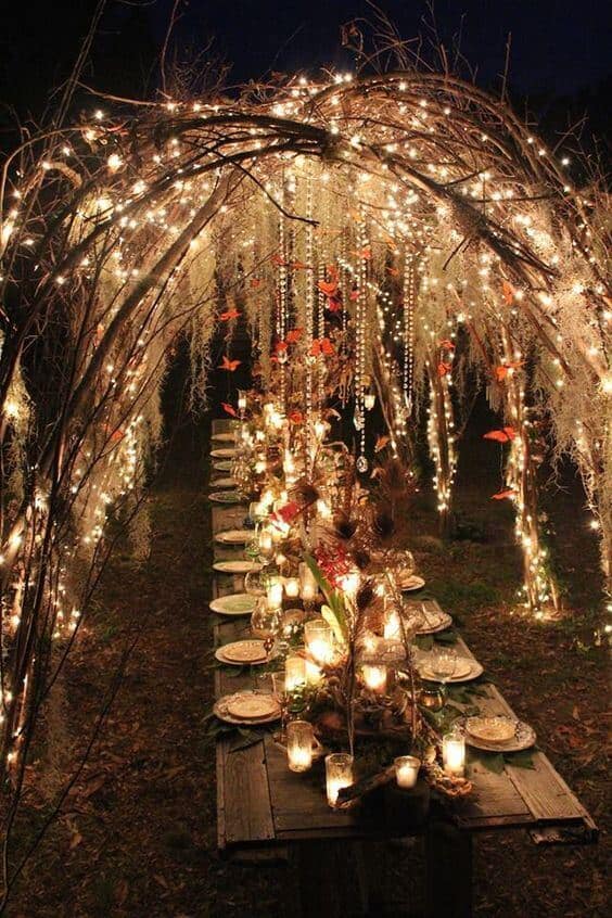 Long rustic wedding table in a shadowy forest, adorned with cascading fairy lights, crystals, and candles under a vine arch.