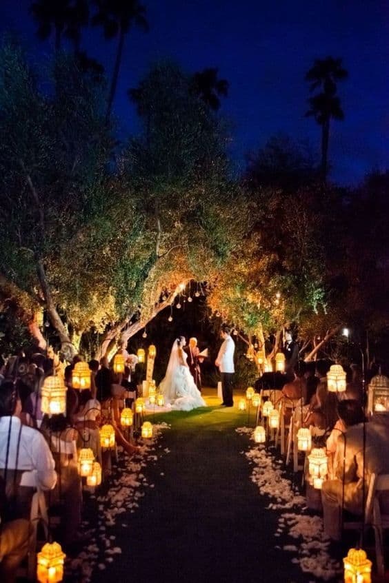 Intimate outdoor night wedding beneath a canopy of glowing lanterns and fairy lights, with a couple exchanging vows along a petal-lined aisle.