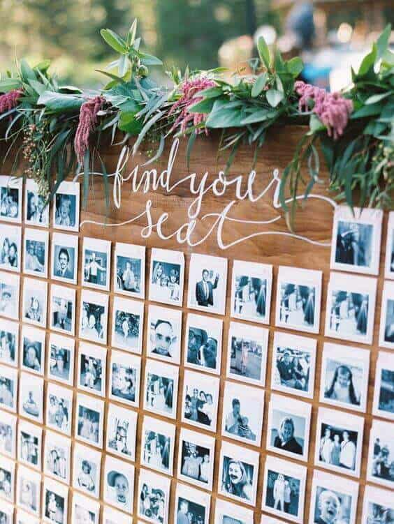 Wooden wedding seating chart featuring white calligraphy "find your seat," adorned with greenery and amaranthus garland, plus Polaroid guest photos.