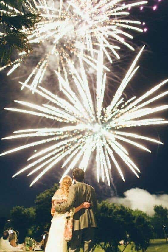 Couple embracing under a midnight sky as white-gold fireworks burst above their outdoor wedding reception, creating a romantic atmosphere.