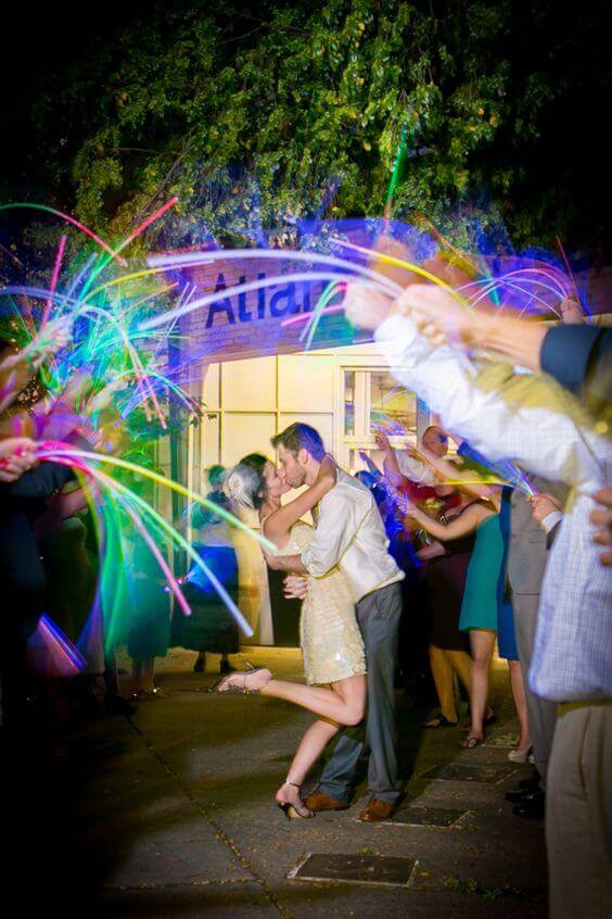 Newlyweds share a dip kiss during a vibrant glow-stick send-off, surrounded by guests waving neon lights at a night wedding.