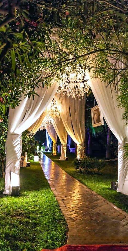 Enchanting night wedding garden walkway with draped ivory arches, crystal chandeliers, and warm lighting creating a romantic atmosphere.