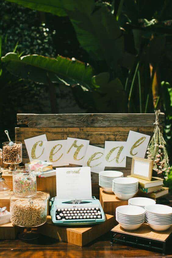 37 Morning Wedding Ideas filled with Sunshine color pallets 25 A rustic breakfast station in a garden with a wooden backdrop, gold glitter "Cereal" sign, jars of colorful cereals, and stacked white bowls.