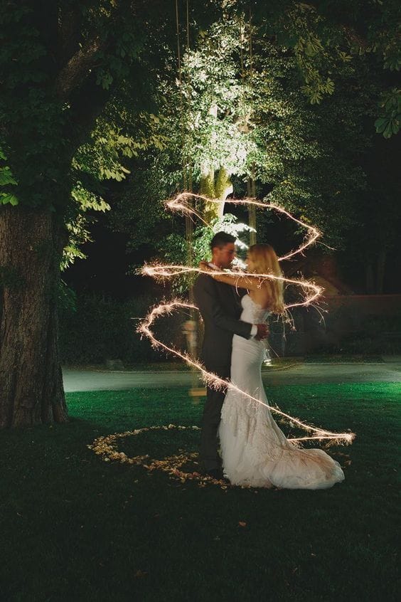 Couple in wedding attire embracing beneath a tree, surrounded by a heart of petals, with a sparkler trail illuminating the night.