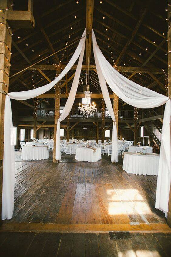 Warm wooden barn transformed into a wedding reception with cascading white chiffon drapes, a crystal chandelier, and round tables dressed in white linens.