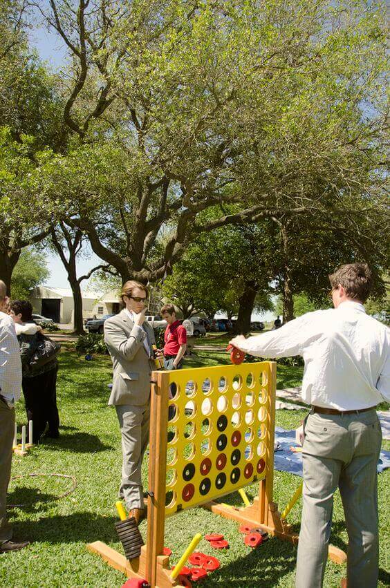 37 Morning Wedding Ideas filled with Sunshine color pallets 26 Guests in light gray suits play giant Connect Four on a sunny lawn, surrounded by mature trees, during a relaxed morning wedding celebration.