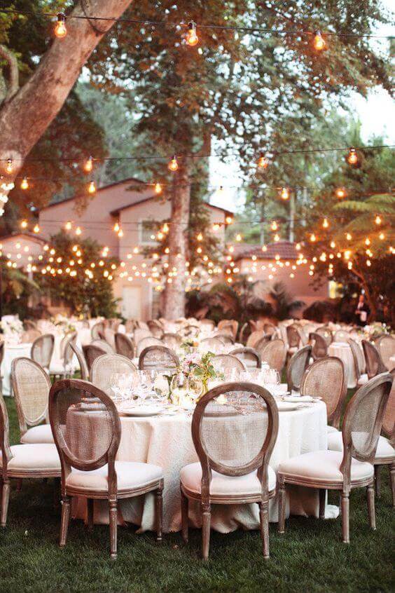A romantic outdoor wedding reception at dusk, featuring round ivory-draped tables, vintage louis chairs, and glowing bistro lights.