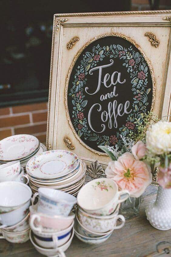 37 Morning Wedding Ideas filled with Sunshine color pallets 27 Vintage-inspired wedding tea and coffee station featuring an ornate chalkboard sign, mismatched floral china, and fresh blooms on a rustic table.