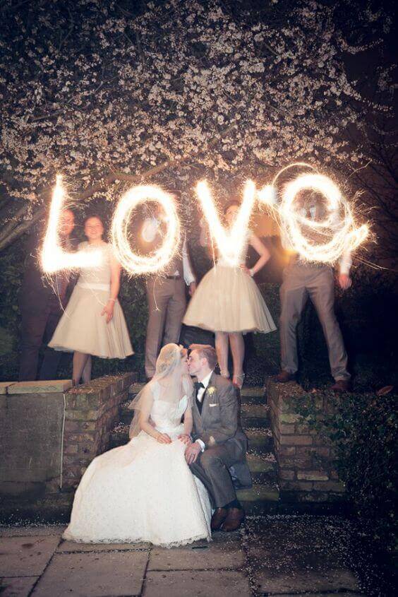 Bride and groom share a kiss on stone steps at night, while attendants create sparkler lettering of "LOVE" under a blooming tree.