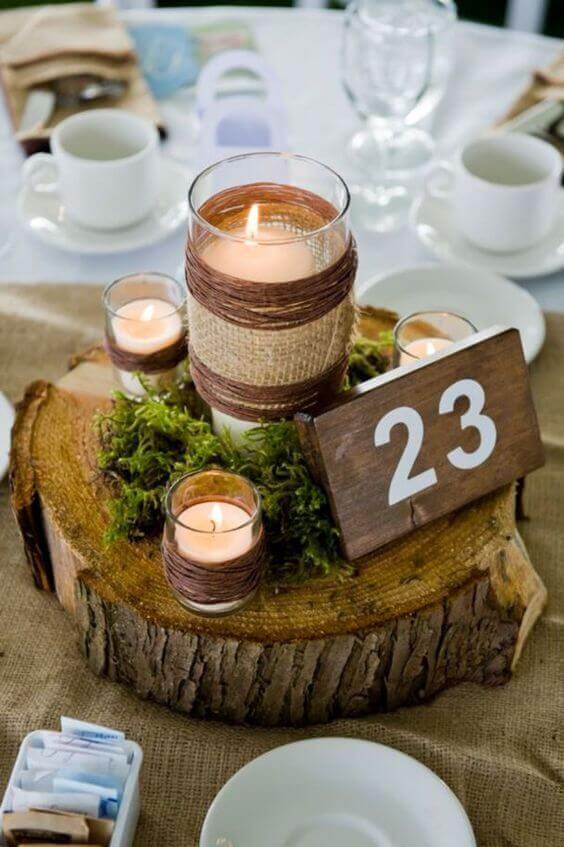 Rustic wedding centerpiece on a wood slice with twine-wrapped candles, green moss, and a table number 23 among white tableware.