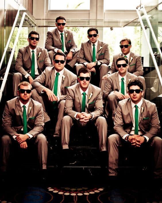 31 amazing wedding groomsmen suits suggestions 26 Nine groomsmen in taupe suits with emerald ties and white boutonnieres pose on a glossy black staircase, exuding modern-urban elegance.