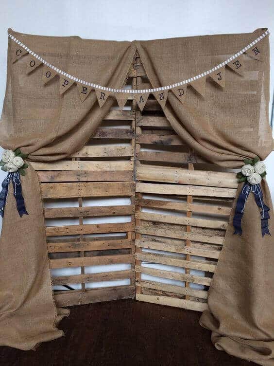 Rustic burlap wedding backdrop featuring weathered wooden pallets, draped burlap, and navy ribbon tiebacks, ideal for barn weddings.