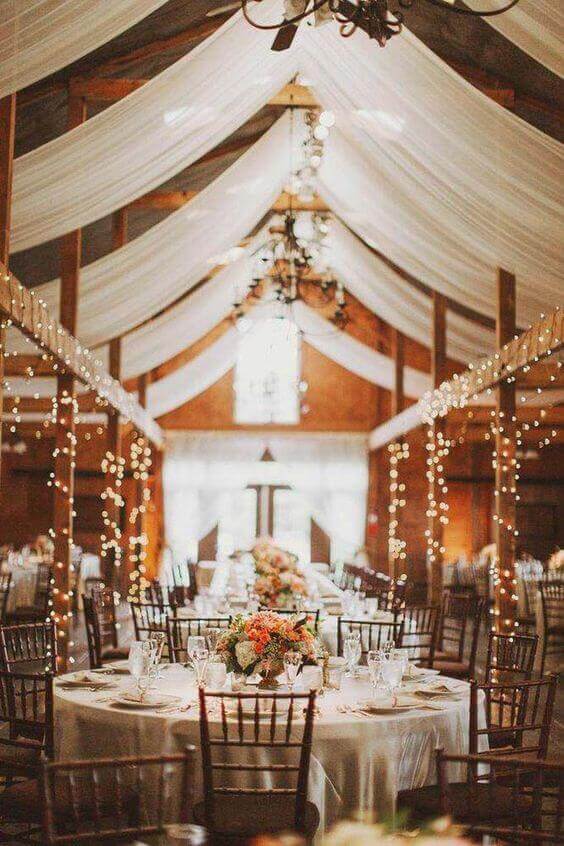 Rustic barn reception with ivory draping, fairy lights, and lush flower centerpieces. Intimate atmosphere for a cozy wedding celebration.