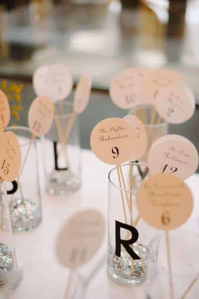 Chic wedding escort card display with blush cards on wooden skewers in clear glass cylinders, elegantly guiding guests to their seats.