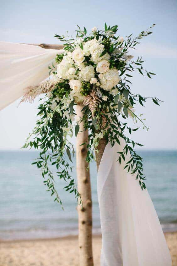 37 Morning Wedding Ideas filled with Sunshine color pallets 29 Coastal wedding arch adorned with flowing white chiffon and lush greenery, set against a serene beach backdrop, capturing a romantic vibe.