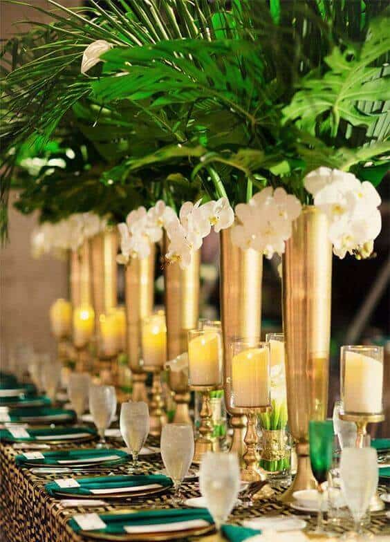 Opulent tropical glam wedding tablescape featuring tall gold vases, white orchids, and flickering candlelight, set for an elegant reception.