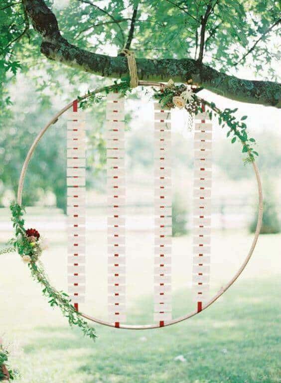 Romantic boho wedding seating display: a large hoop hangs from a tree, adorned with cascading ivory cards and greenery, creating an elegant focal point.