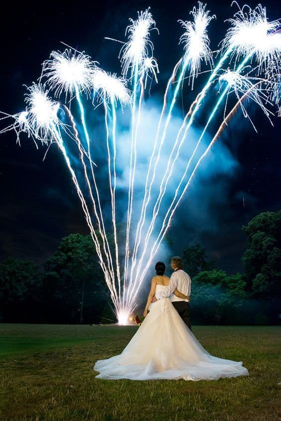 Newlyweds embrace on a dark lawn as icy-white fireworks light up the sky, creating a romantic and magical night wedding moment.