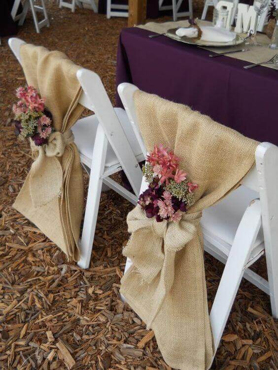 White folding chairs adorned with burlap sashes and pink-and-berry wildflower posies, set beside a plum-linen table on rustic wood-chip ground.