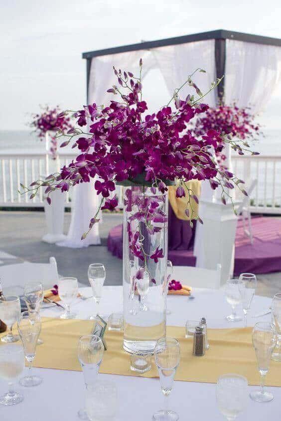 Tall glass vase filled with fuchsia-purple dendrobium orchids as a wedding table centerpiece on a seaside deck, with a white canopy behind.