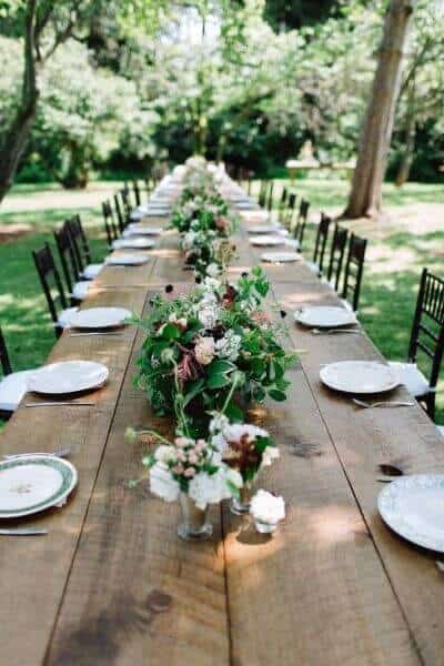 37 Morning Wedding Ideas filled with Sunshine color pallets 33 Sun-dappled garden wedding reception featuring a long farmhouse table, low floral runner, mismatched vintage china, and dark wooden chairs.