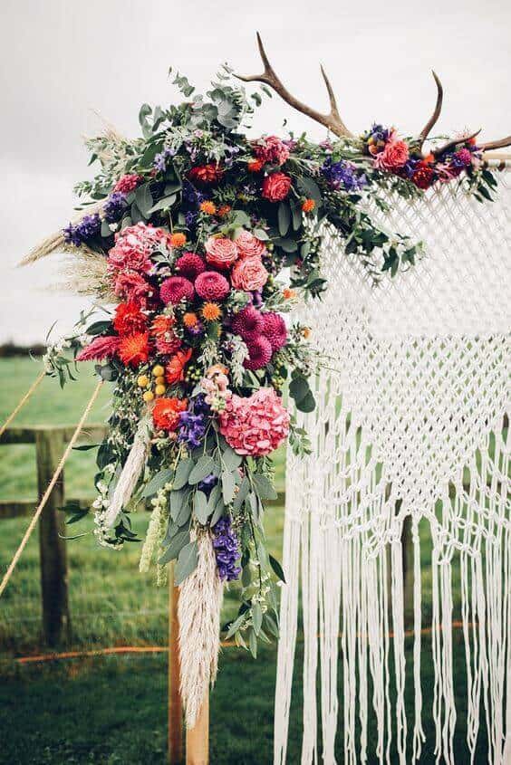 Boho wedding arch featuring macramé and vibrant jewel-tone florals, set in a rustic meadow with a soft overcast sky.