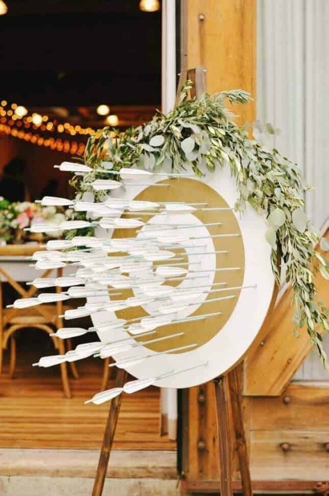 Rustic-chic wedding seating display featuring a circular target board with gold rings, arrows as escort cards, and lush greenery in a barn setting.