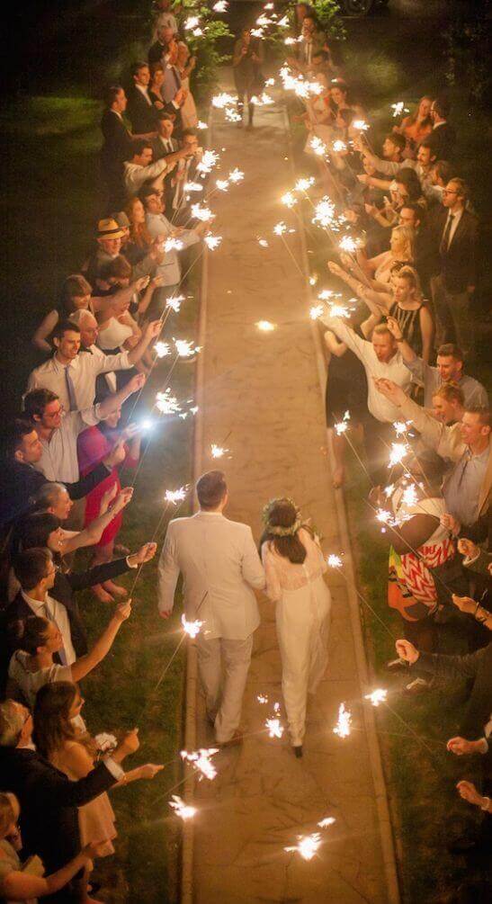 Just-married couple walking hand-in-hand down a sparkler-lit stone aisle at night, surrounded by cheering guests in a romantic garden setting.