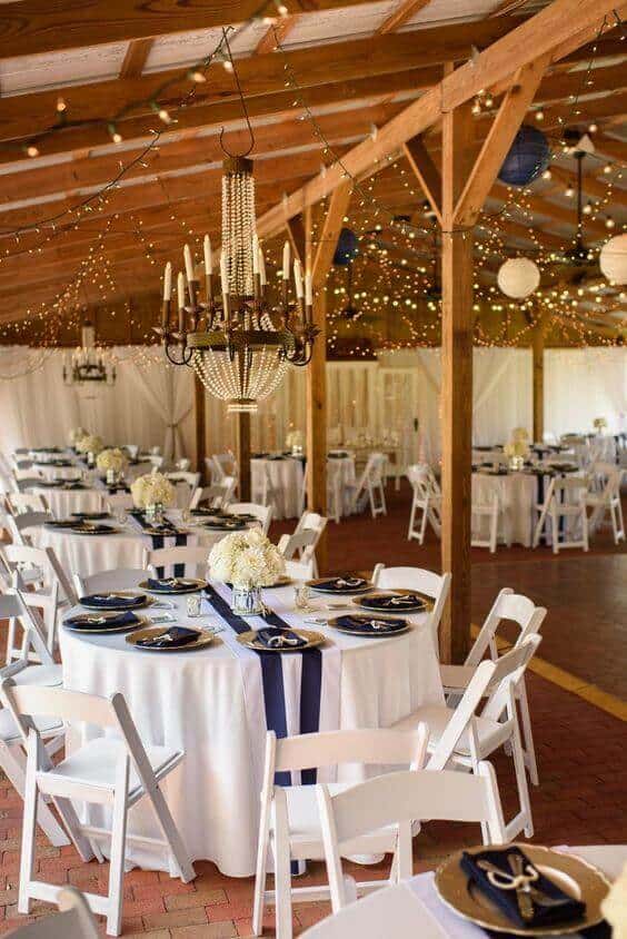 Rustic barn wedding reception featuring navy-and-gold tables, pearl chandelier, and fairy lights, creating a warm, inviting ambiance.