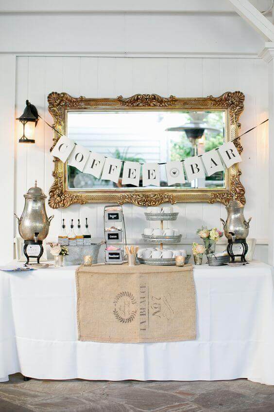 37 Morning Wedding Ideas filled with Sunshine color pallets 35 A rustic wedding coffee bar featuring a white-linen table, silver urns, tiered pastry stand, and a gold-framed mirror with "COFFEE BAR" bunting.